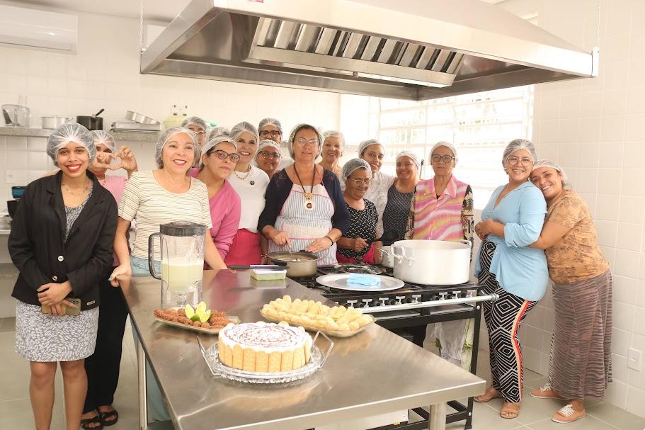 Grupo de mulheres à frente de mesa com doces e salgados. #paratodosverem