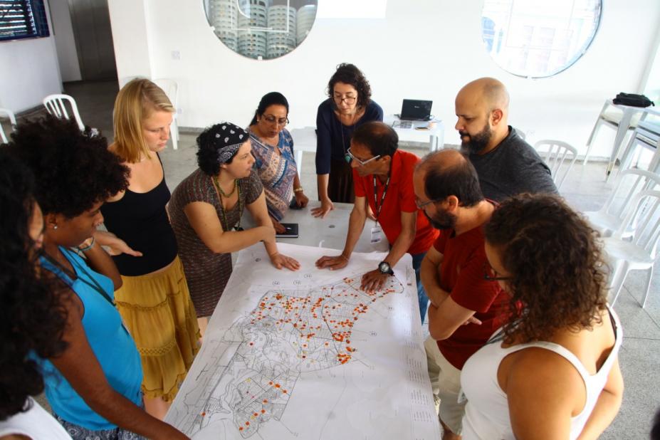 Grupo de pessoas em pé em torno de uma mesa observa mapa da Cidade. #Pracegover