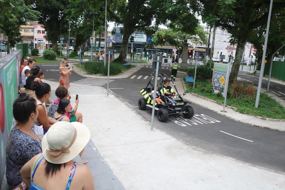 adultos próximos a um muro observam criança dentro de um minibug que passa na pista da praça. #paratodosverem 
