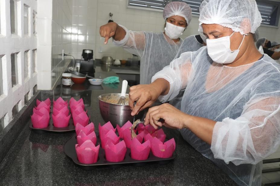 mulher usando touca, avental e máscara está depositando massa de bolo em forminhas sobre uma bandeja. #paratodosverem