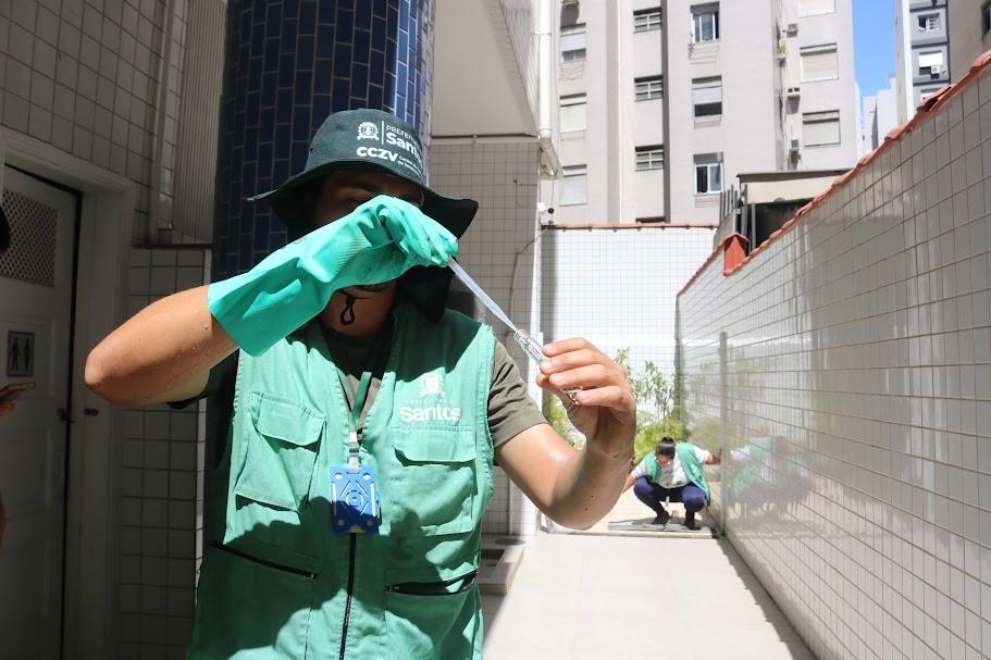 agente uniformazido e com bone, usando luva em uma das mãos segura tubo de ennsaio com larva. Ao fundo, outra agente está fazendo uma coleta. #paratodosverem 