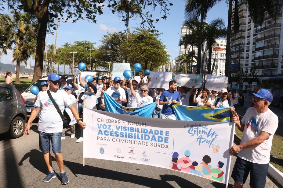 pessoas uniformizadas carregam faixa e cartazes na orla. #paratodosverem