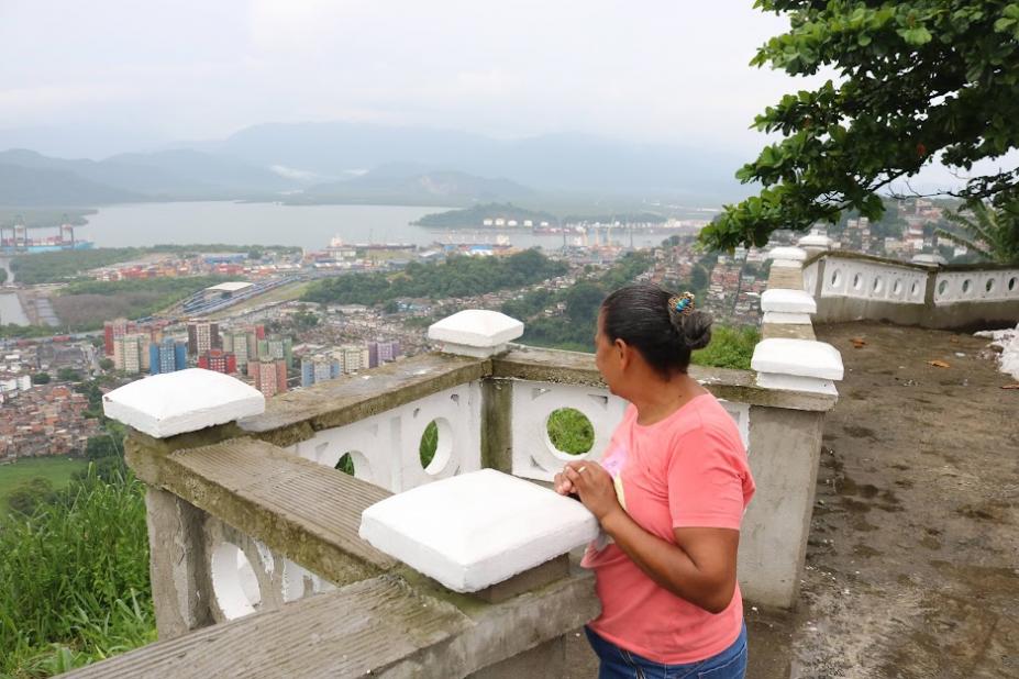 mulher está atrás de mureta no alto de morro olhando para a parte de baixo da cidade, com diversas construções, e o porto ao fundo. #paratodosverem