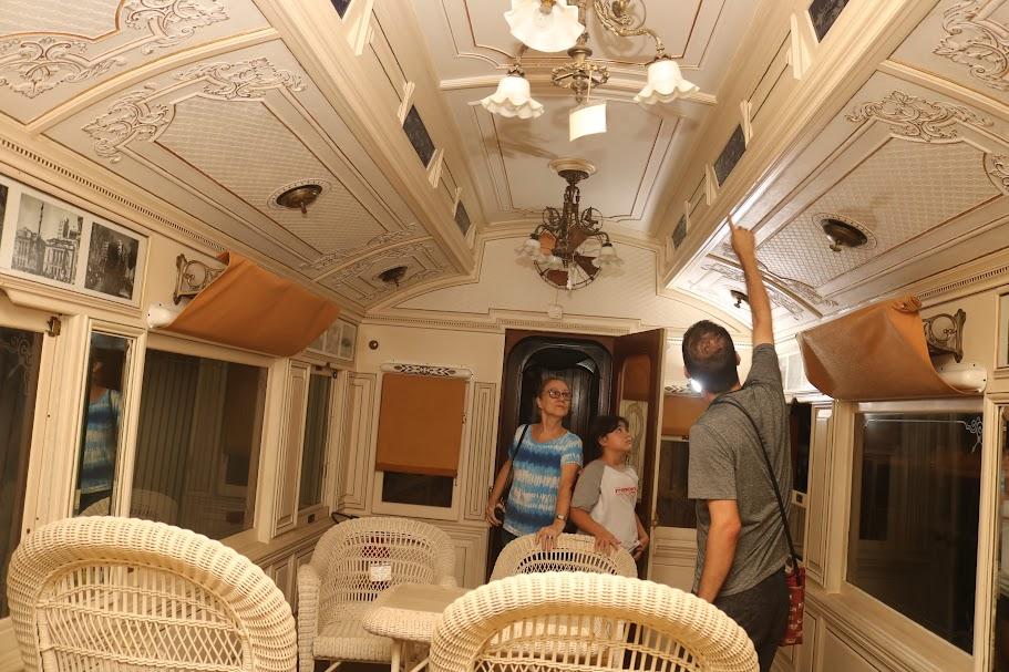 Três pessoas estão dentro de um carro ferroviário. duas são visitantes e um é o guia que aponta para um detalhe no teto. #paratodosverem 