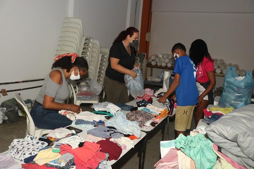 Várias pessoas realizam triagem de roupas que estão em cima da mesa. #paratodosverem