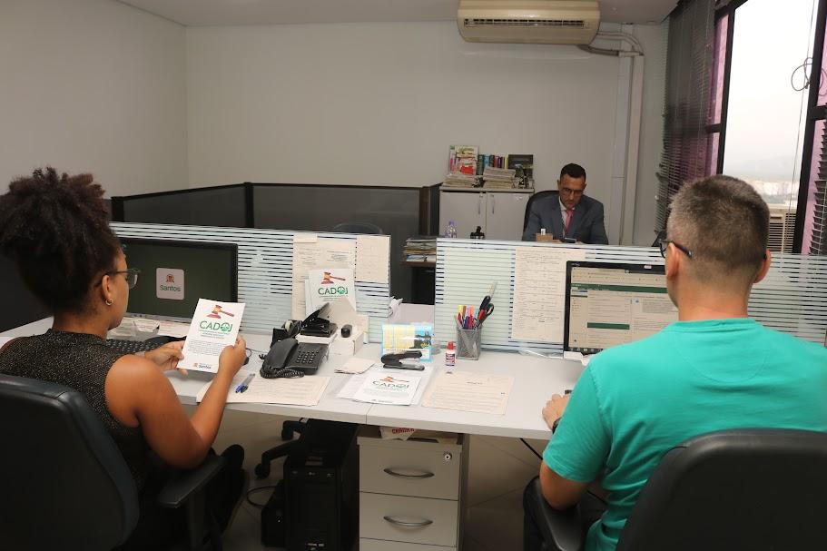 dua pessoas sentadas de costas para a foto diante de computador. Mulher, à esquerda, segura folheto do cadoj. #paratodosverem 
