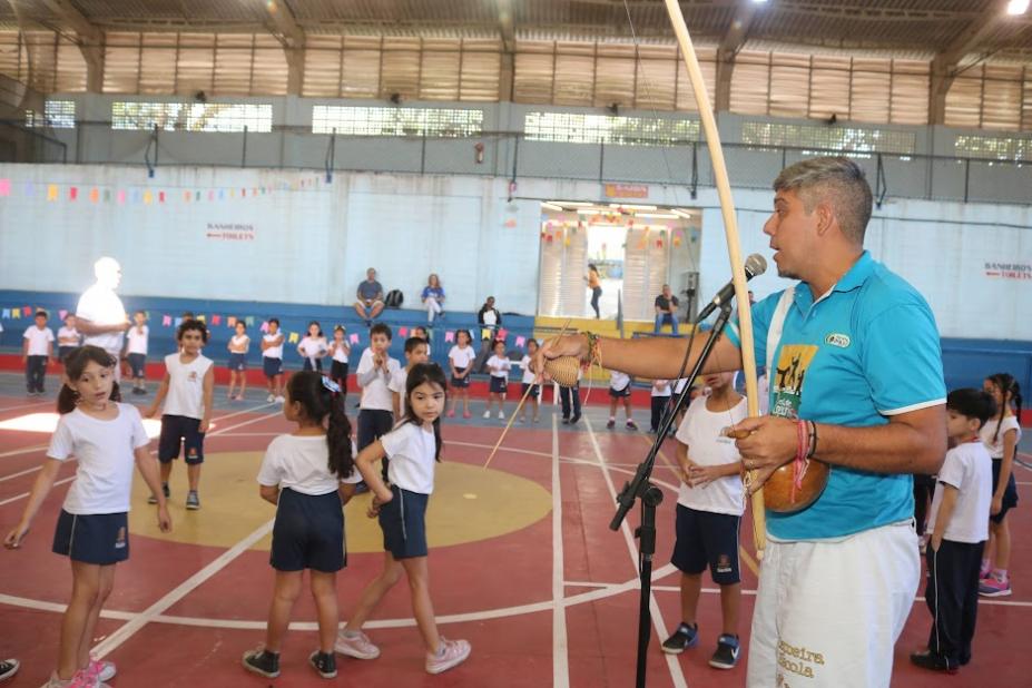 homem toca berimbau e canta próximo a microfone. Crianças estão na quadra da escola. #paratodosverem 