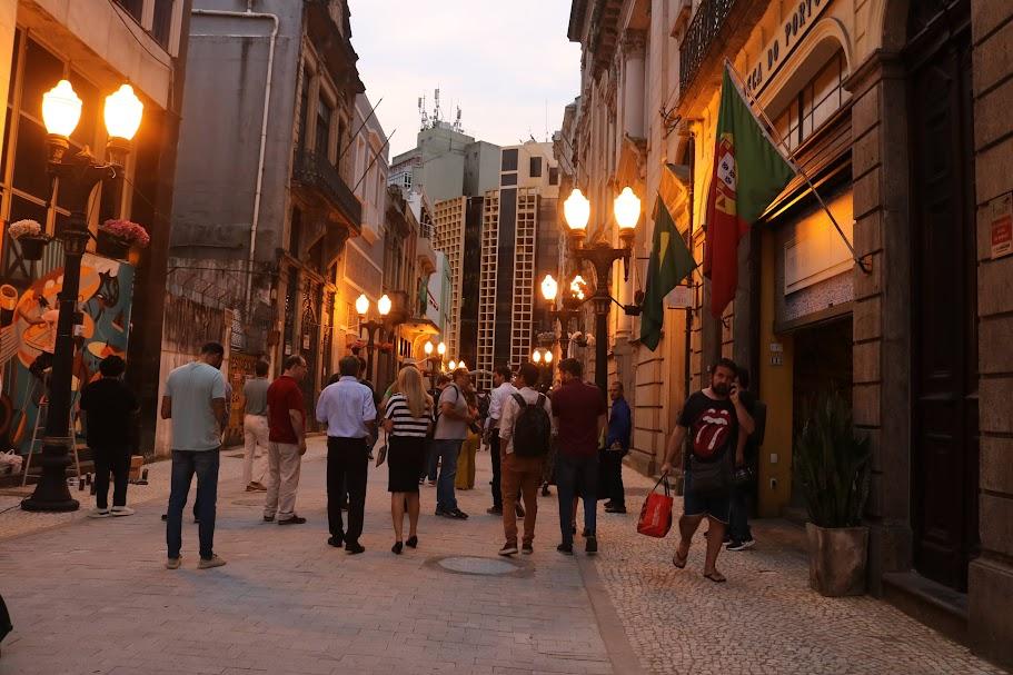 pessoas circulam em calçadão de rua ao cair da tarde. Luminárias estão acesas. #paratodosverem