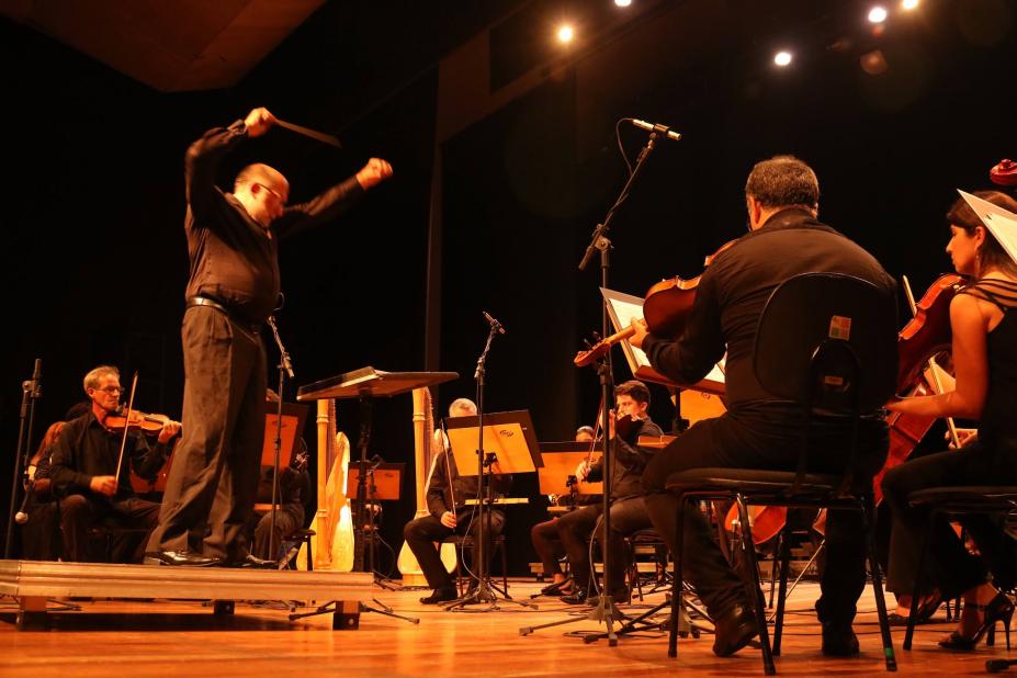 maestro em pé rege orquestra em palco. #paratodosverem