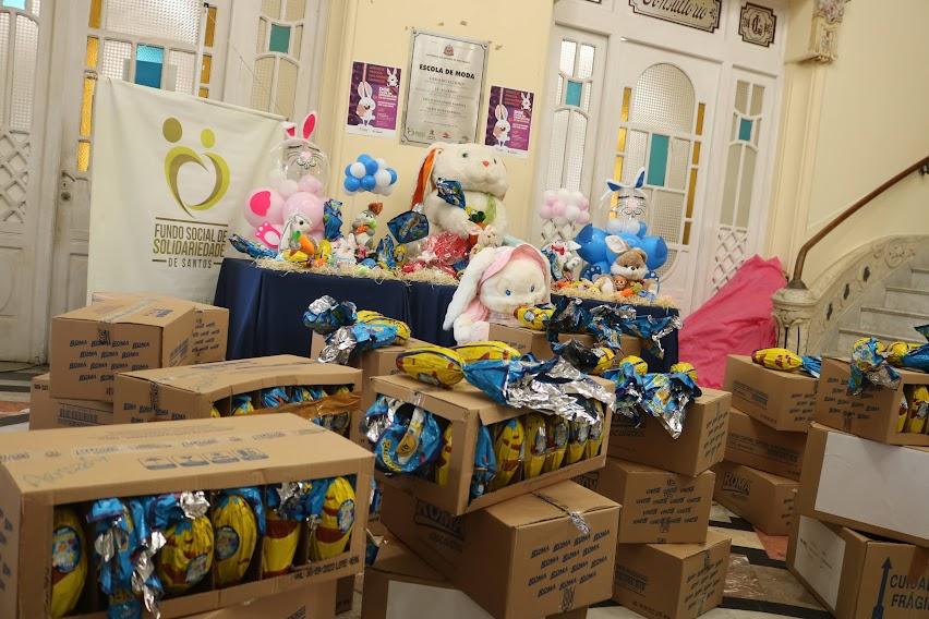 ovos de páscoa dentro e fora de caixas estão expostos em salão. Sobre uma mesa e sobre uma caixa há coelhinhos de pelúcia. #paratodosverem 