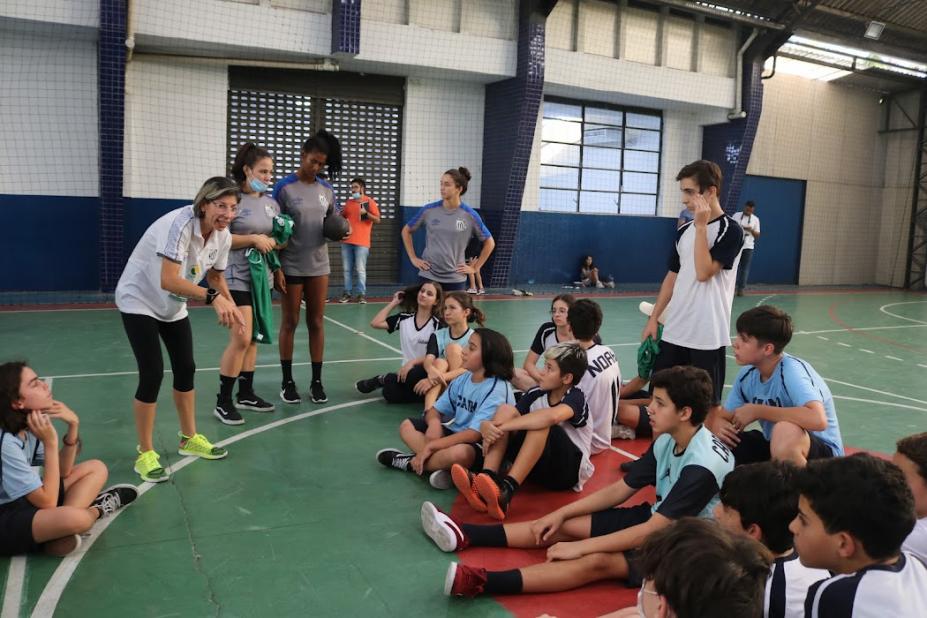 tecnica e atletas conversam com alunos sentados em quadra #paratodosverem