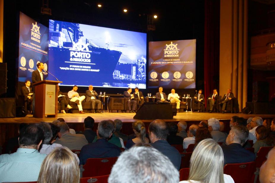 Palco de teatro com autoridades sentadas e um homem falando em uma tribuna. Em primeiro plano está a plateia, de costas para a imagem. #Pracegover