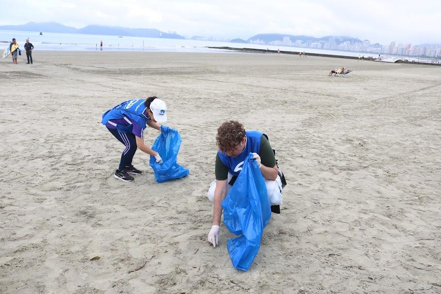 duas pessoas estão na faixa de areia coletando resíduos e levando-os a um saco plástico. #paratodosverem 