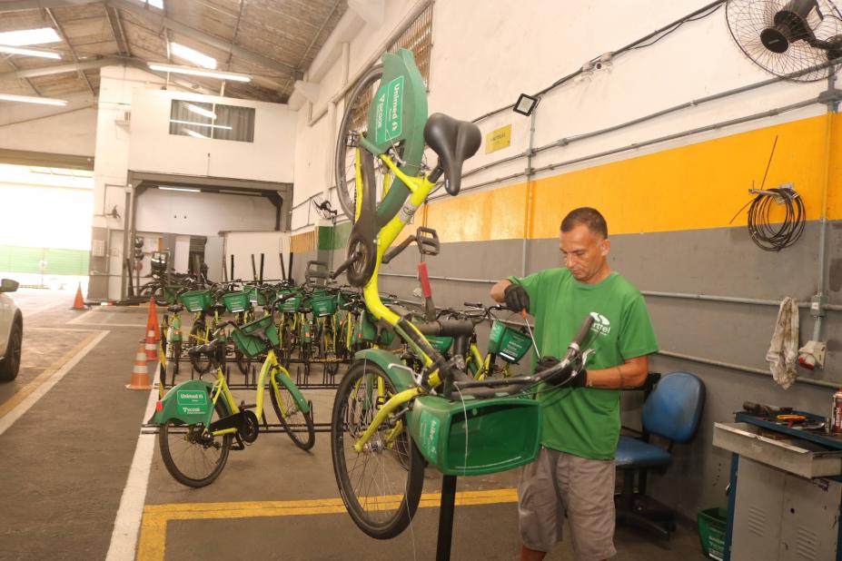 Homem conserta bicicleta. há bikes ao fundo do galpão. #paratodosverem
