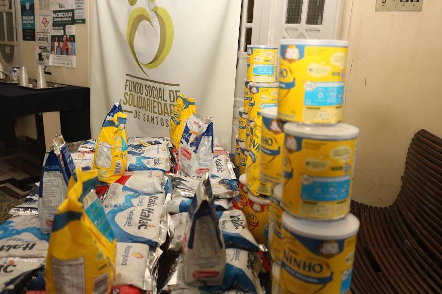 latas e pacotes de leite em pó sobre uma mesa, com banner do fundo social ao fundo. #paratodosverem