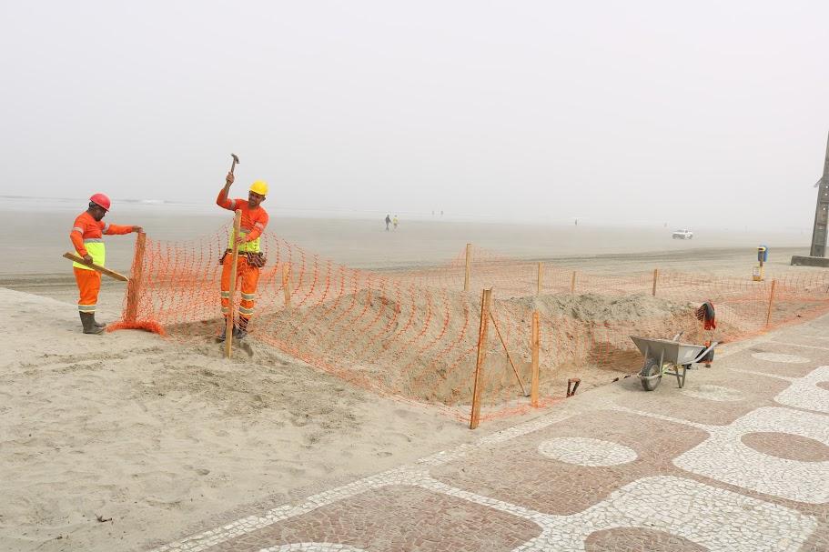 dois homens estão cercando com lona plástica perfurada trecho de areia na praia, junto ao calçadão. #paratodosverem 