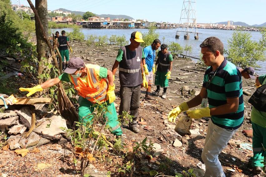 homens estão recolhendo detritos em área de mangue. Todos usam luvas. #paratodosverem 