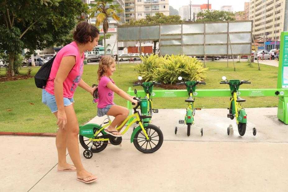 criança pedala bicicleta com a mãe em pé ao lado. #paratodosverem