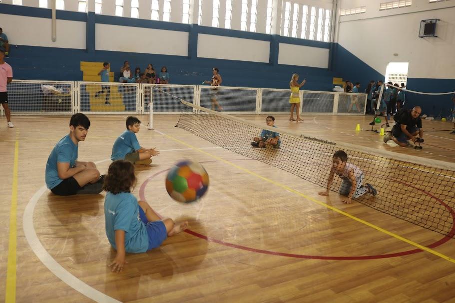 crianças jogam bolam em quadra. Todas estão sentadas. Há uma rede dividindo a quadra para o vôlei sentado. #paratodosverem 