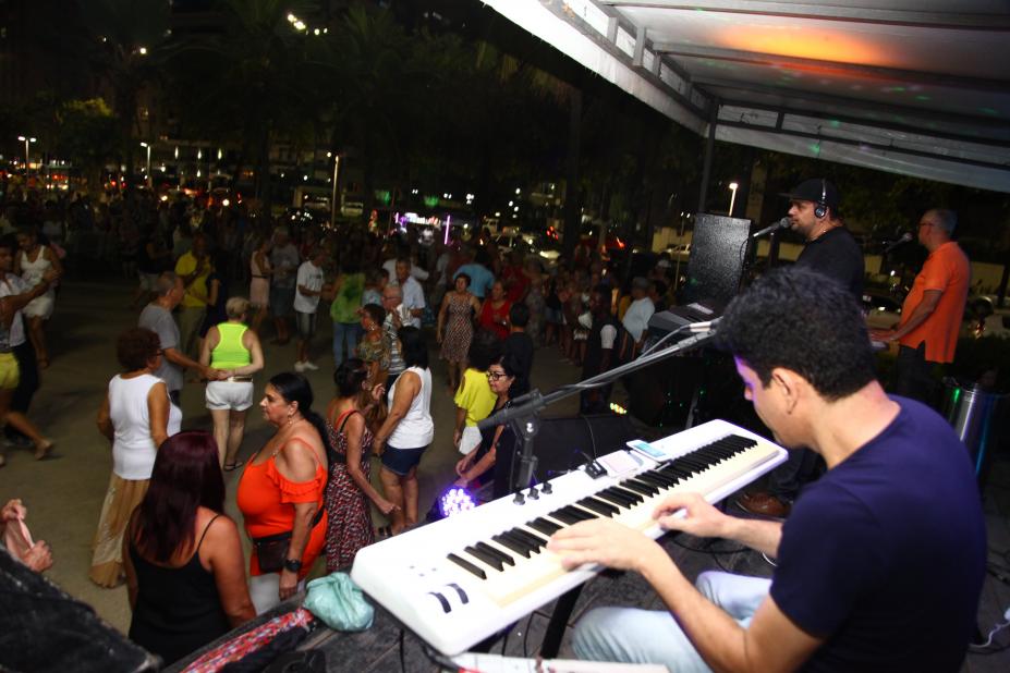 Em foto noturna, homem toca teclado sobre palco enquanto dezenas de pessoas dançam em área do calçadão da praia