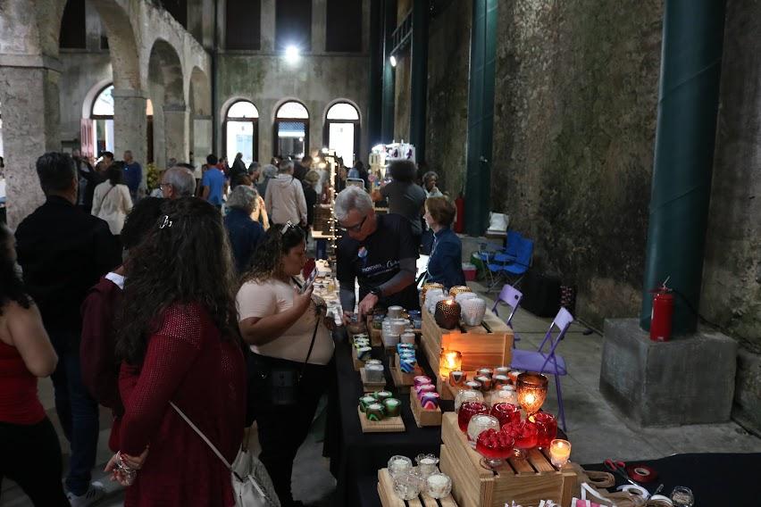 estande com exposição de velas em primeiro plano e pessoas circulando em um salão. #paratodosverem 