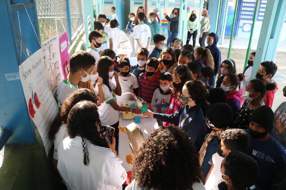 crianças cercam mesa com outros estudantes mostrando objetos #paratodosverem