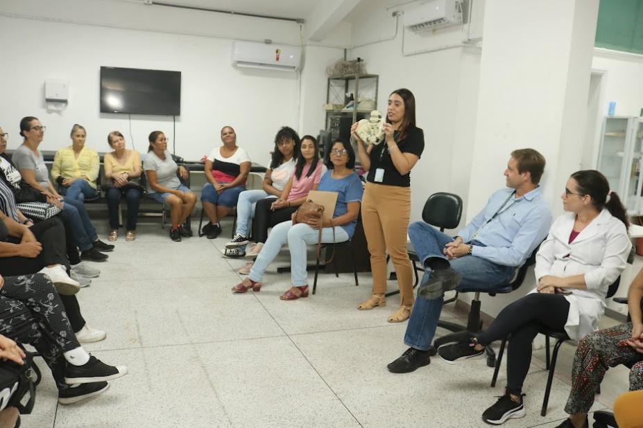 pessoas estão sentadas em círculo assistindo palestra. uma mulher fala em pé ao grupo. #paratodosverem