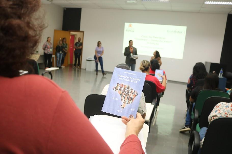 pessoas assistindo palestra. Em primeiro plano, uma mulher, de costas, segura um folheto com instruções para o cuidado com idosos. #paraatodosverem 