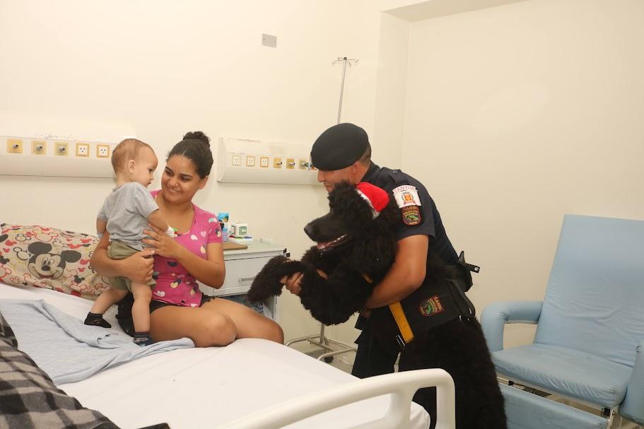 guarda municipal segura cão e aproxima a patinha de uma criança que está no colo da mãe, sentada em leito hospitalar. #paratodosverem 