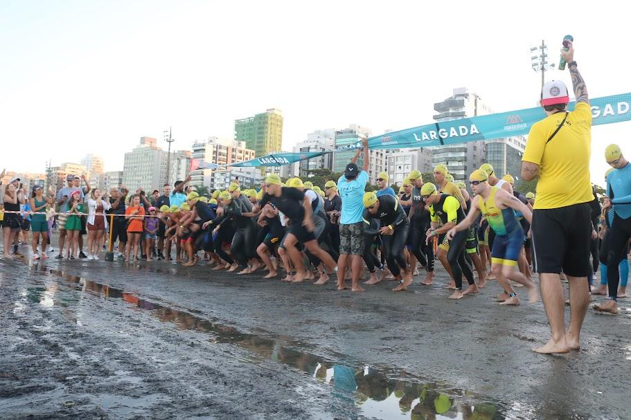 atletas dão largada na prova de natação. Eles estão na beira do mar e começam a correr em direção à água. #paratodosverem 