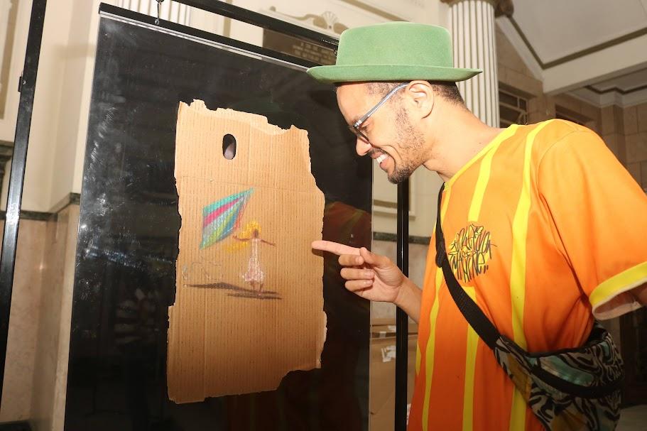 artista aponta para uma de suas obras afixada em display. #paratodosverem 