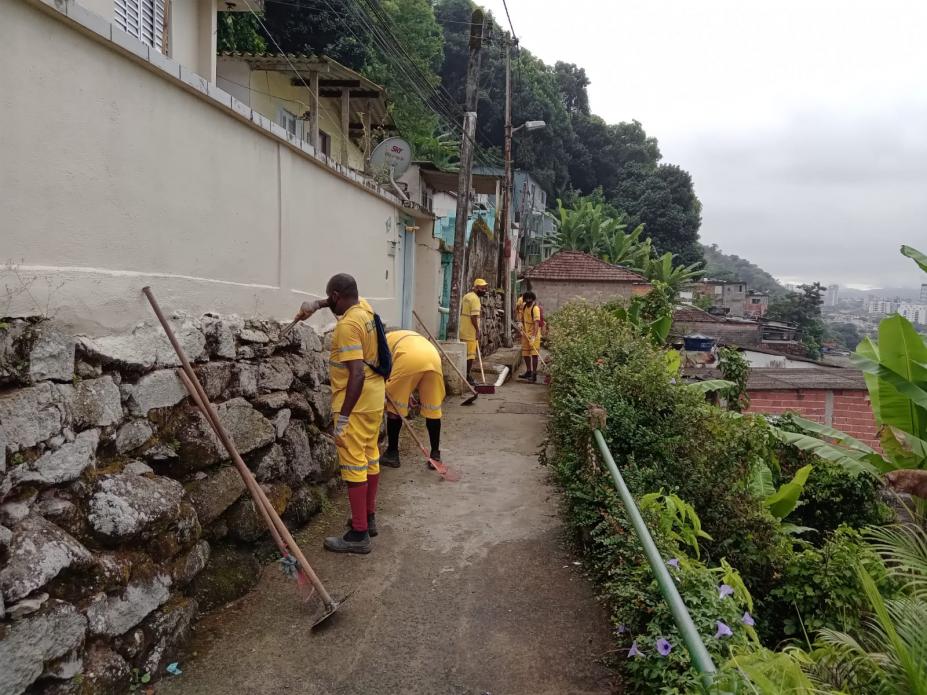 homens uniformizados raspam pedras em muro de contenção em morro. Ao lado direito há vegetação. #paratodosverem