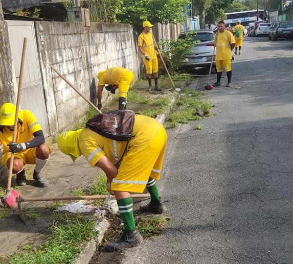 homens uniformizados raspam calçada cheia de mato. #paratodosverem