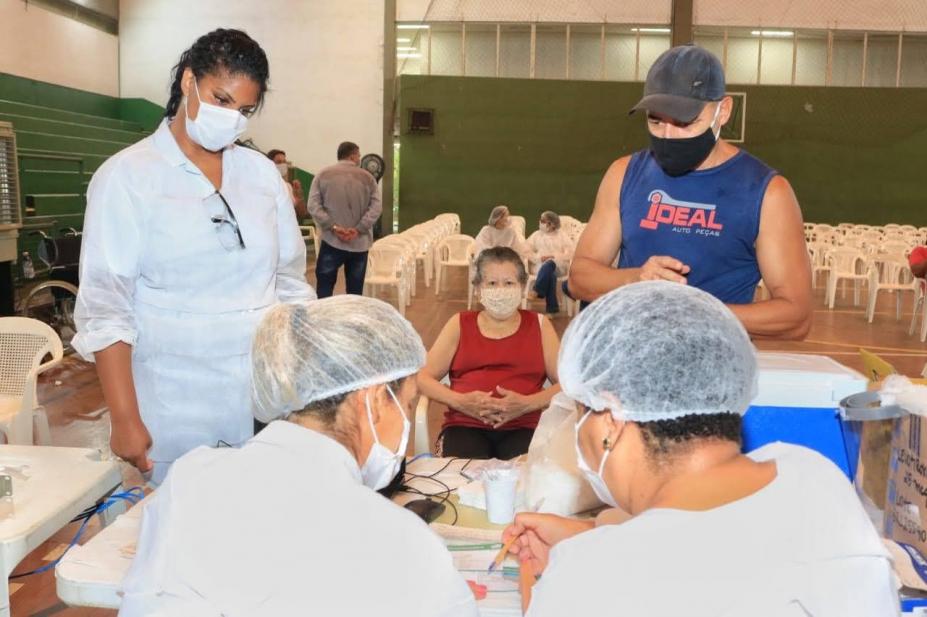 Duas mulheres vestidas de branco estão sentadas, de costas para a imagem. À frente delas, sentada, está uma idosa, aguardando ser atendida para vacinação. A foto foi feita num ginásio esportivo. #Paratodosverem
