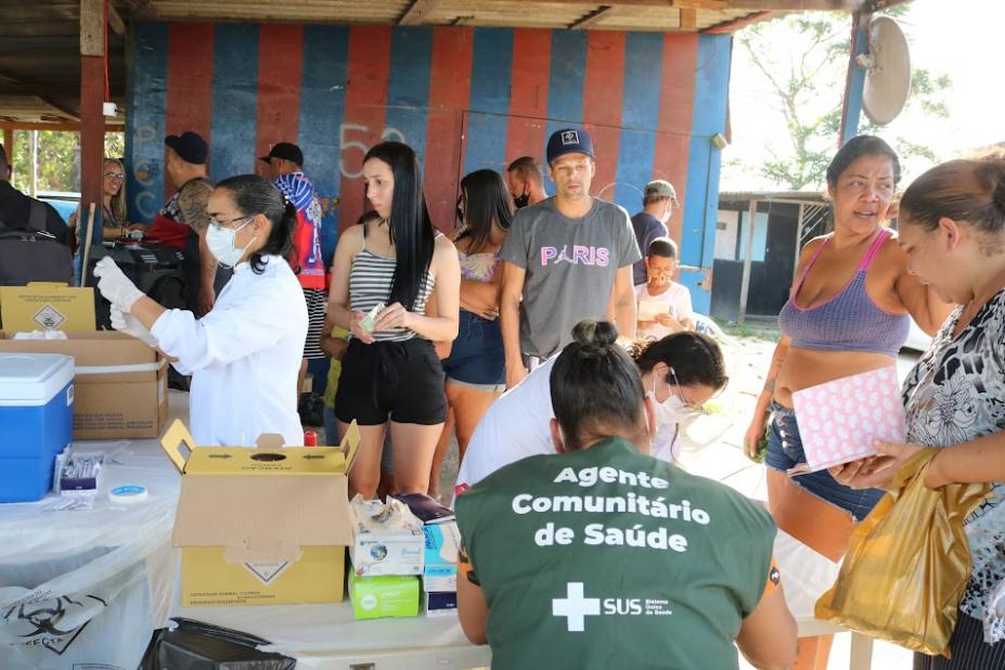 em primeiro plano, mulher usa colete de agente de saúde. Ela está de costas atendendo pessoas que estão sob uma área aberta nas laterais e coberta na parte de cima. Ao fundo, uma mulher prepara uma vacina. #paratodosverem