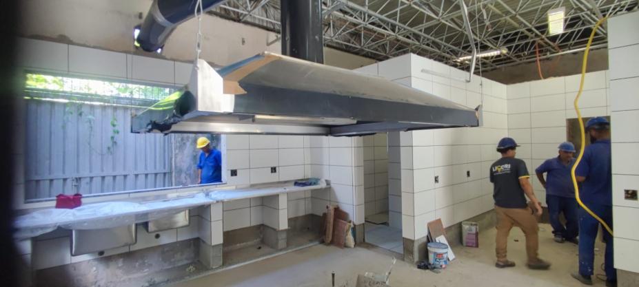 homens trabalham em área que é uma cozinha. Há obras no local. É possível ver a estrutura de uma coifa industrial. #paratodosverem 