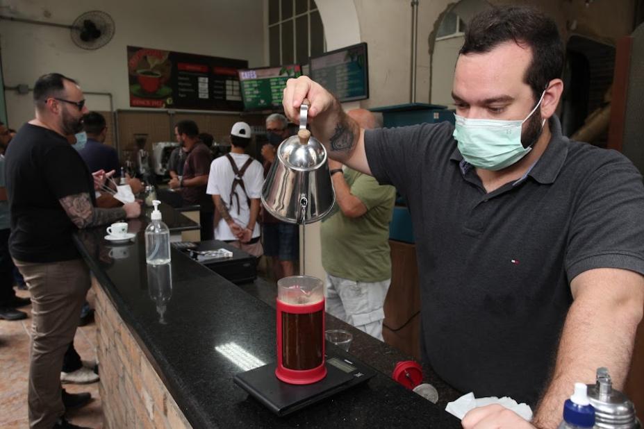 HOMEM SEGURA BULE E DESPEJA ÁGUA EM COPO PREPARANDO UMA BEBIDA DE CAFÉ NUM COPO LONGO. MOVIMENTAÇÃO DE PESSOAS AO FUNDO. #PARATODOSVEREM