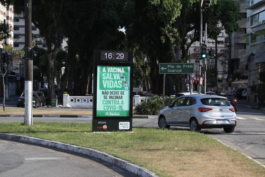 Totem em canteiro central com horário marcando 16h29. O restante do totem tem informação sobre a importância da vacina. Há um carro fazendo curva ao lado. #paratodosverem