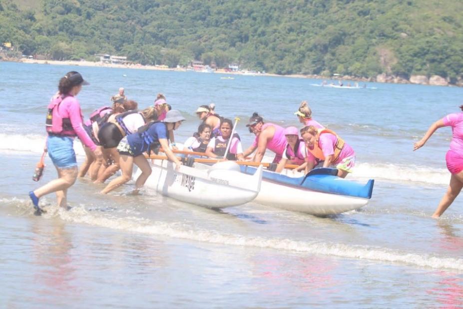 mulheres entram no mar, ainda empurrando a canoa havaiana. Morro ao fundo. #paratodosverem