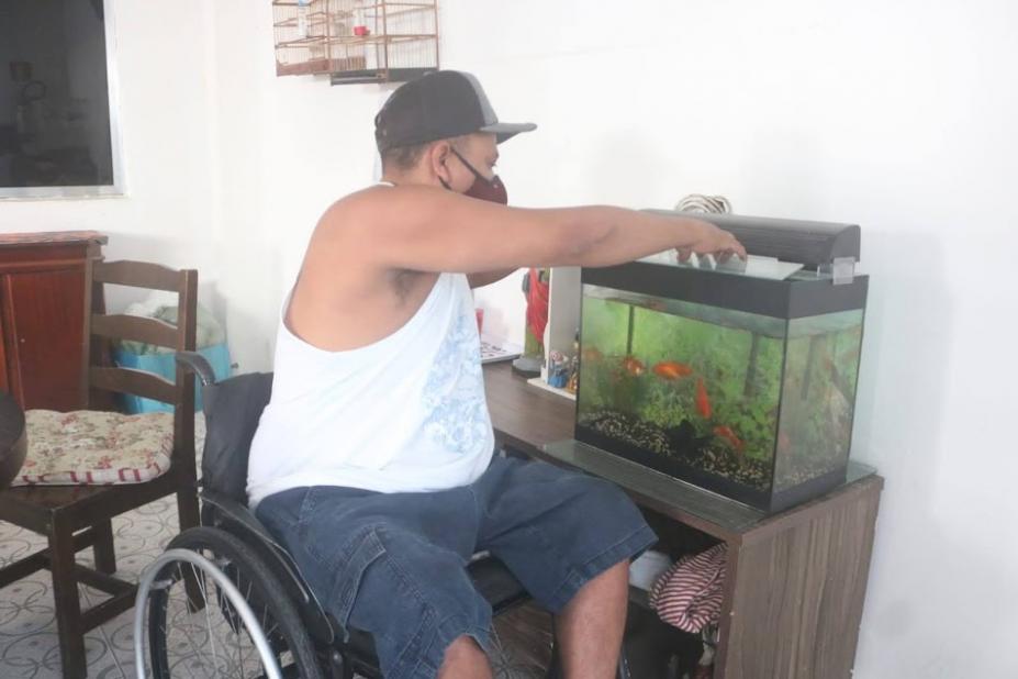 homem está em cadeira de rodas e mexe em aquário que está sobre um móvel. #paratodosverem
