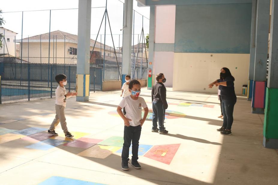 alunos estão em pátio fazendo atividade com monitora. Eles estão afastados. Ao fundo, há uma quadra esportiva cercada. #paratodosverem