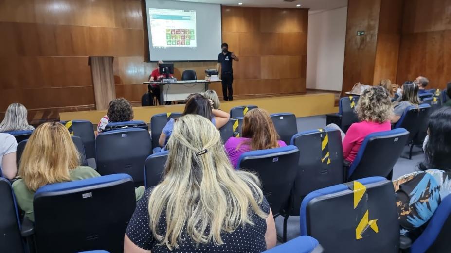 Pessoas sentadas em auditório assistindo palestrante #paratodosverem