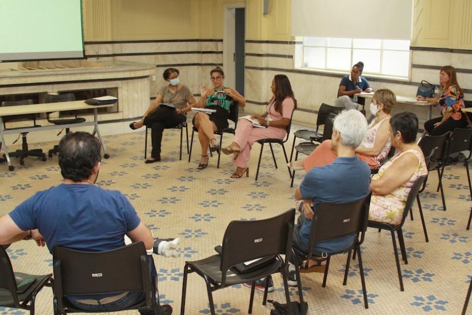 várias pessoas sentadas em roda em uma sala. Uma delas, ao fundo, está falando. #paratodosverem