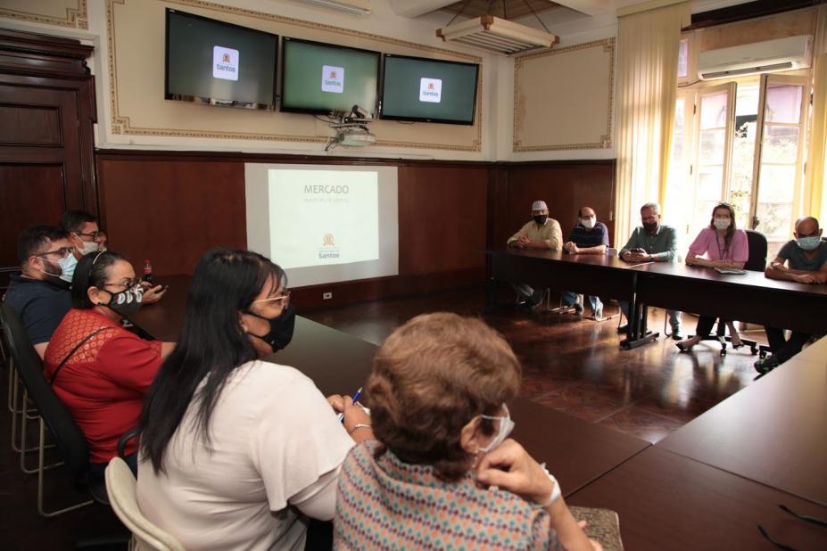 pessoas reunidas em torno de uma mesa em forma de letra U. Ao fundo, três monitores e um telão. #paratodosverem