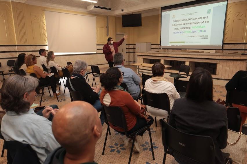 sala de reunião com um homem à frente de todos os que assistem. Ele aponta para um projetor na parede que tem informações sobre o programa Município Verde Azul. #paratodosverem 