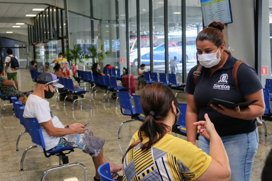 Agente da Ouvidoria conversa com mulher sentada na sala de espera da rodoviária. Há outra pessoas sentadas no mesmo ambiente. #Paratodosverem