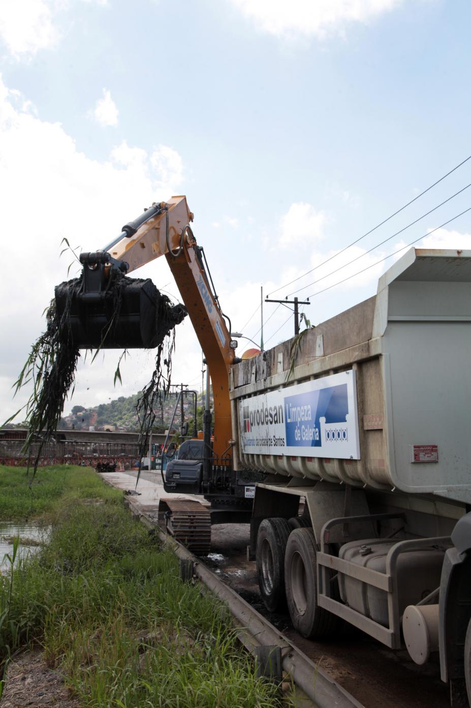 Escavadeira cheia de detritos está elevada em movimento para depositá-los no caminhão que está à frente. #Paratodosverem
