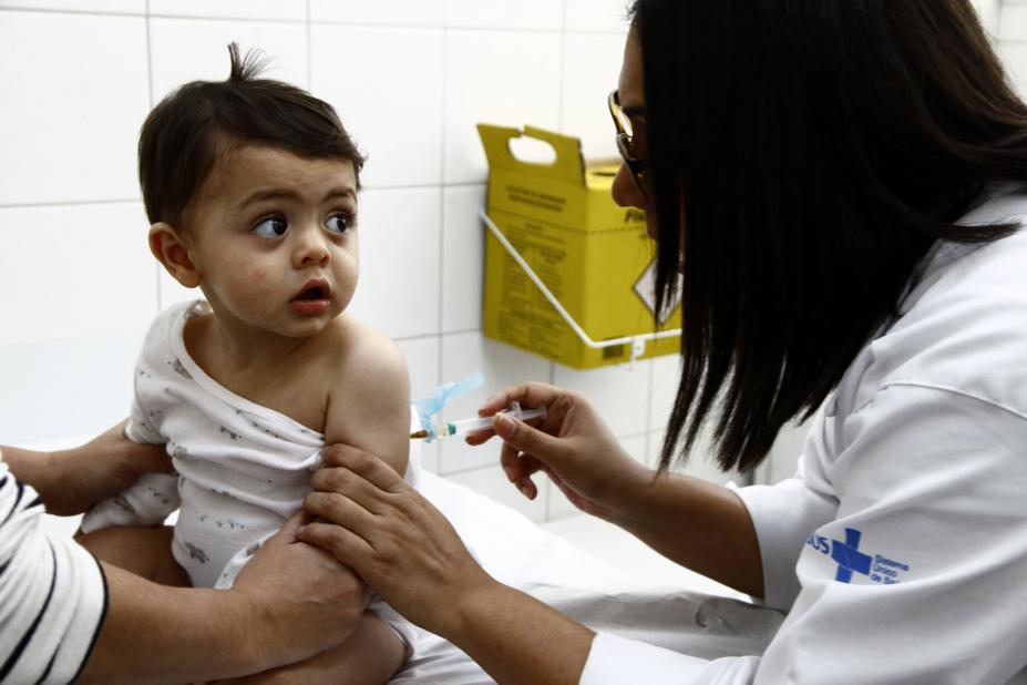 Bebê é vacinado por mulher vestida com avental branco. A criança está sentada em uma maca e uma pessoa segura seu corpo com as duas mãos. #Pracegover