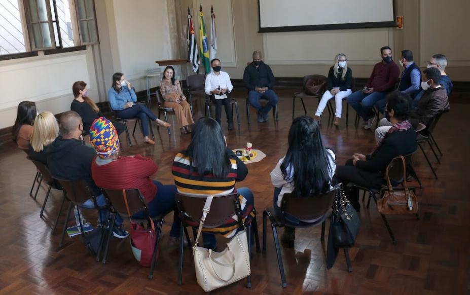 Em uma sala, várias pessoas sentadas em um circulo conversando. #paratodosverem