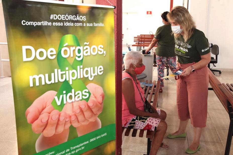 mulher conversa com idosa sentada, ao lado cartaz sobre doação de órgão. #paratodosverem